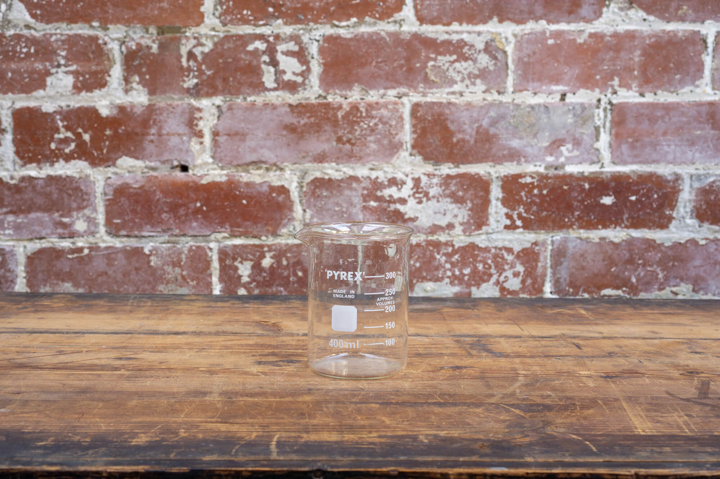 Photo shows a selection of vintage glass laboratory vessels on a table. The background is a red rustic brick wall.