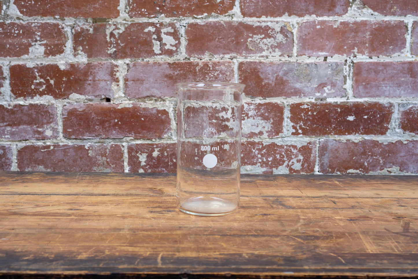 Photo shows a selection of vintage glass laboratory vessels on a table. The background is a red rustic brick wall.