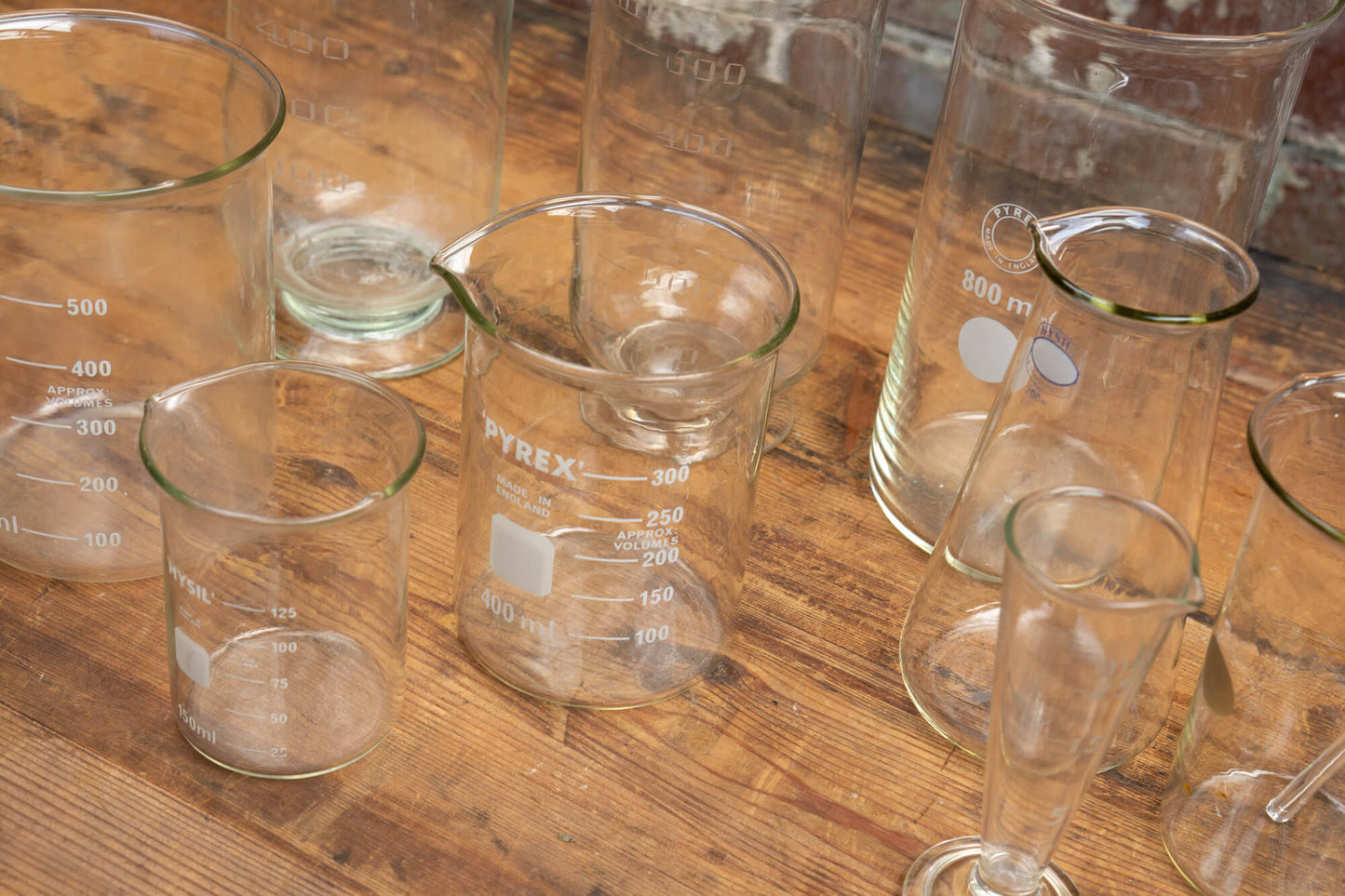 Photo shows a selection of vintage glass laboratory vessels on a table. The background is a red rustic brick wall.