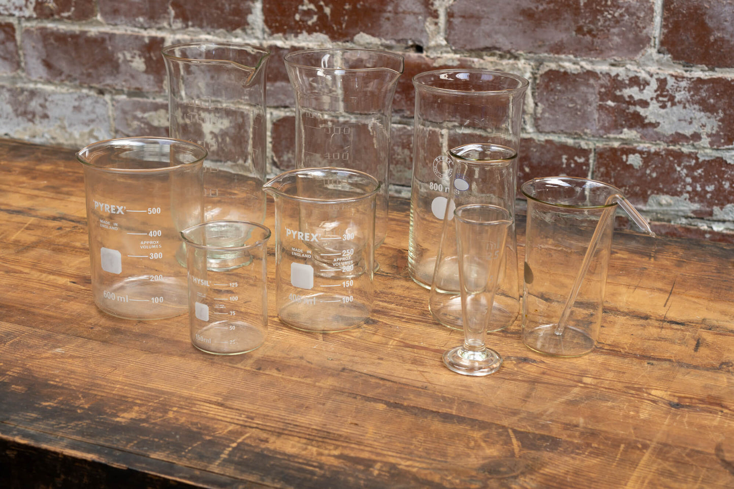 Photo shows a selection of vintage glass laboratory vessels on a table. The background is a red rustic brick wall.