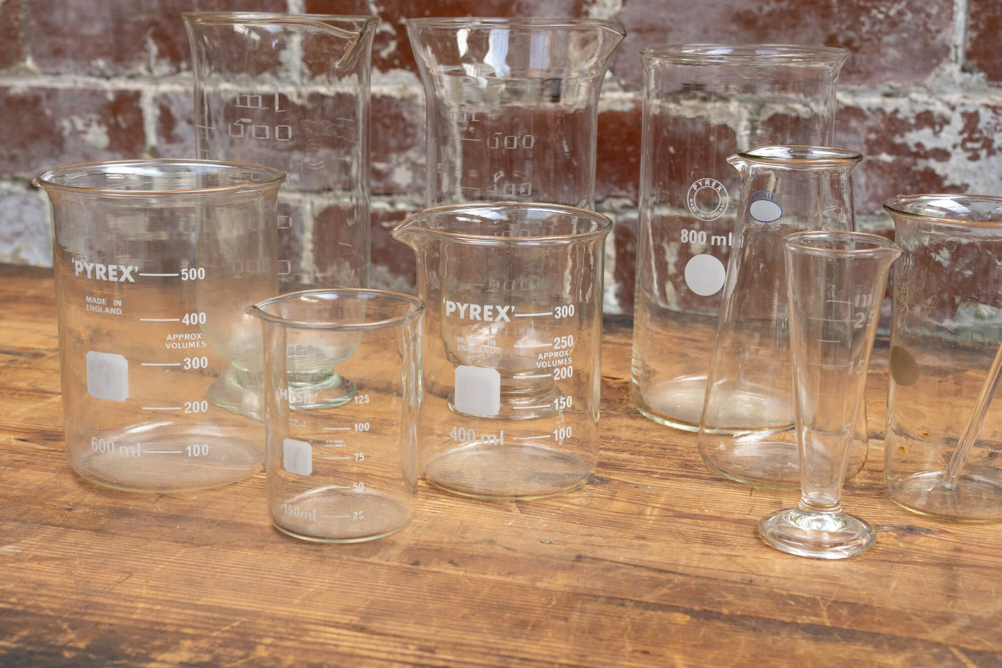 Photo shows a selection of vintage glass laboratory vessels on a table. The background is a red rustic brick wall.