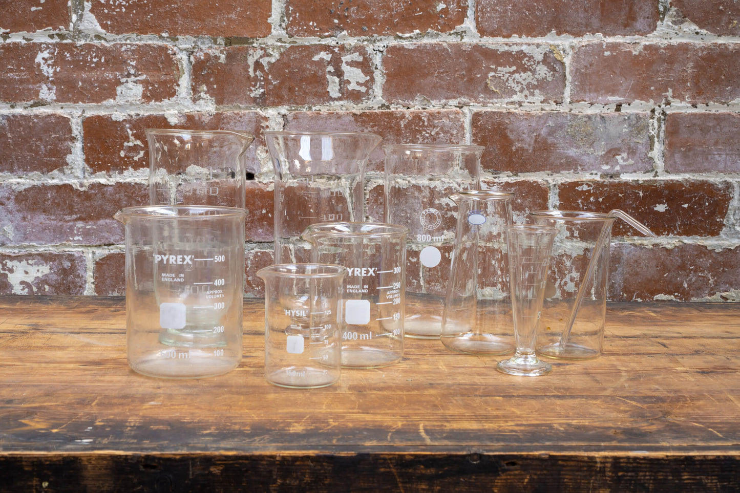 Photo shows a selection of vintage glass laboratory vessels on a table. The background is a red rustic brick wall.