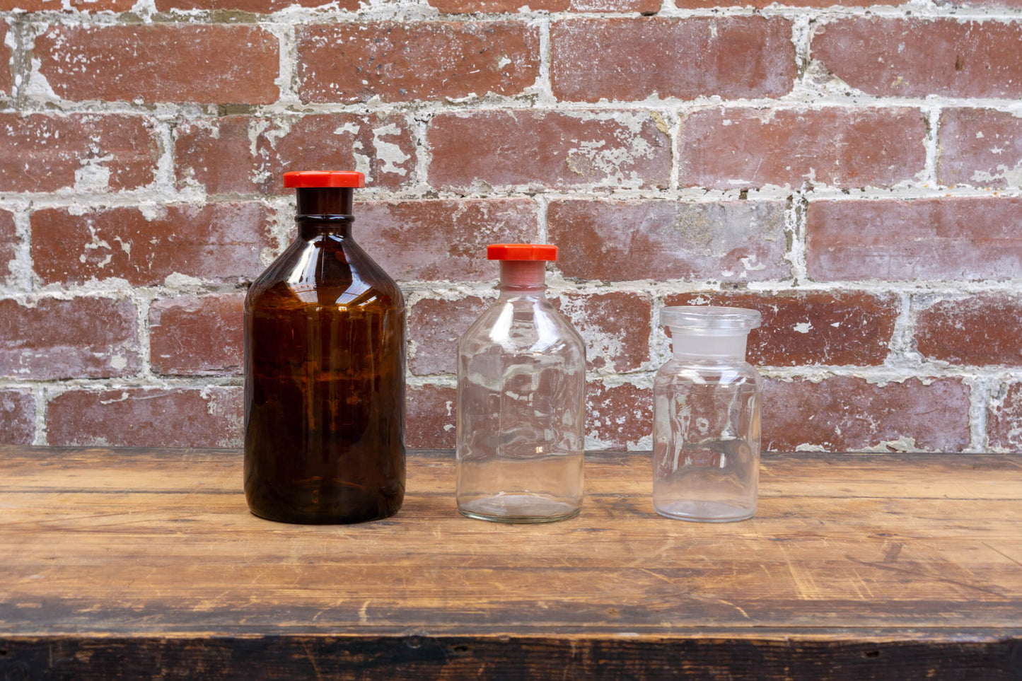 Photo shows a selection of vintage apothecary style glass science bottles atop a wood table. The background is a red rustic brick wall.