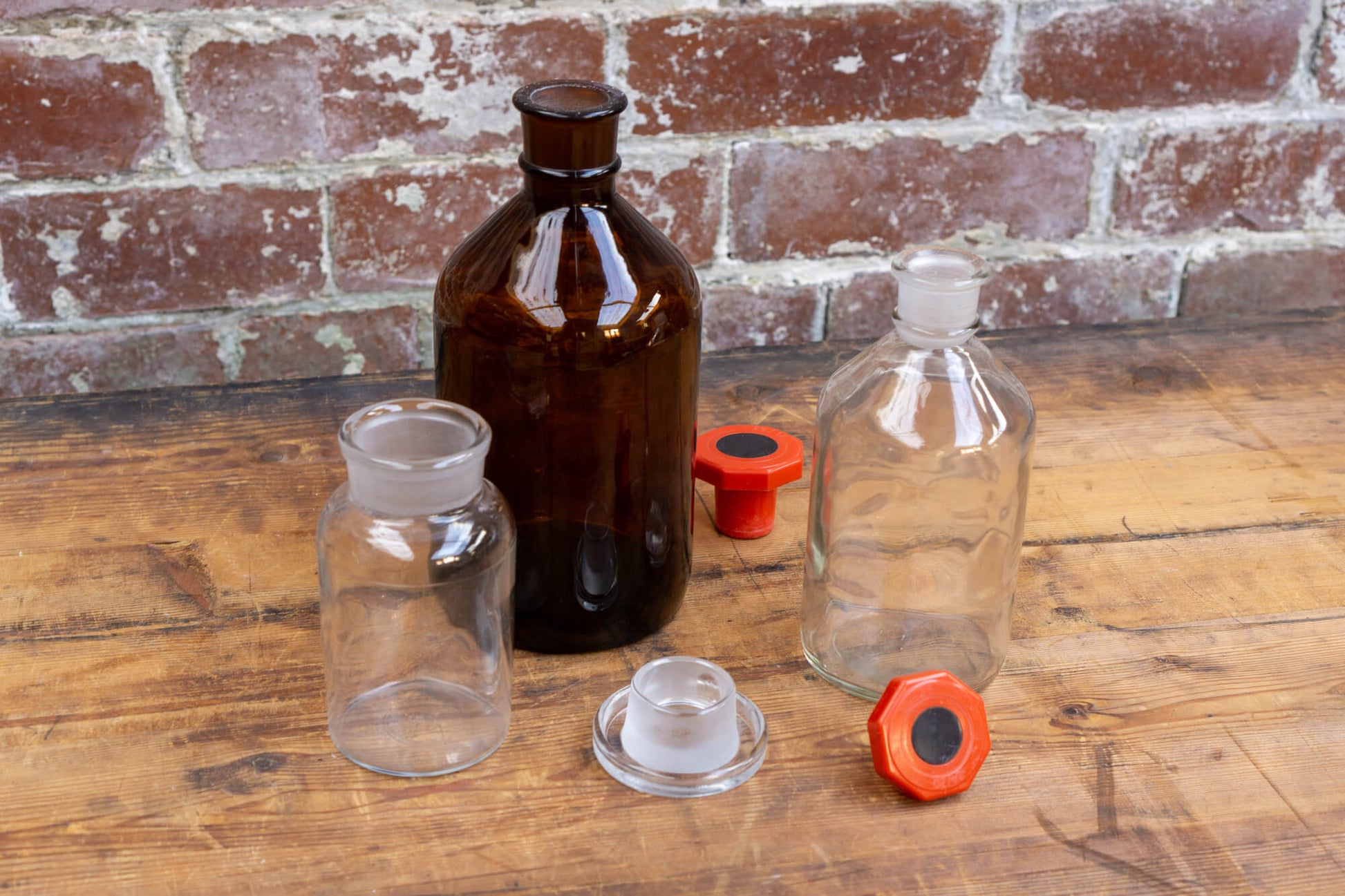 Photo shows a selection of vintage apothecary style glass science bottles atop a wood table. The background is a red rustic brick wall.
