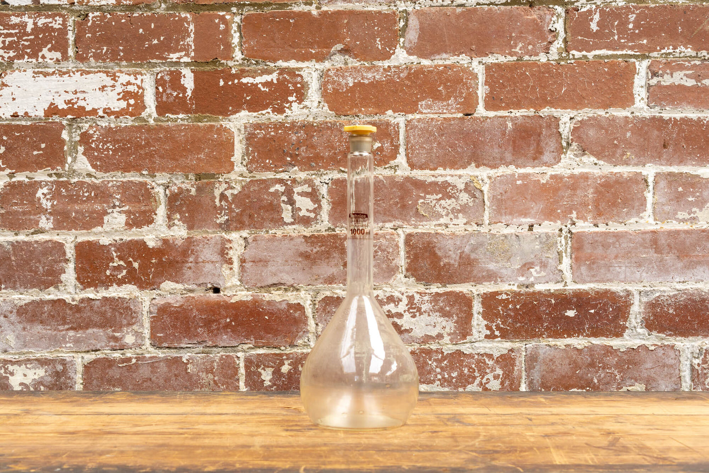 Photo shows a selection of vintage glass laboratory vessels on a table. The background is a red rustic brick wall.