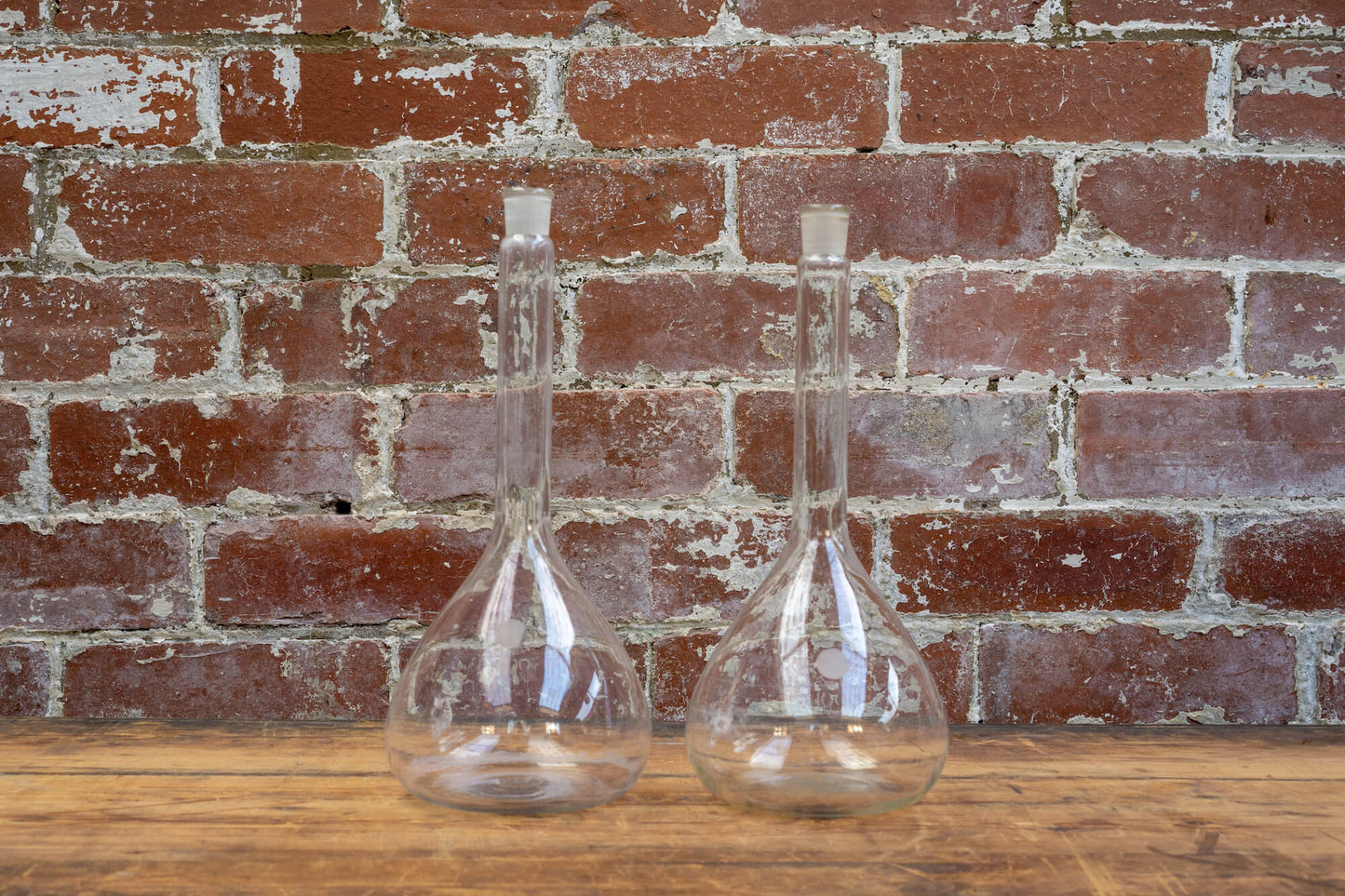 Photo shows a selection of vintage glass laboratory vessels on a table. The background is a red rustic brick wall.