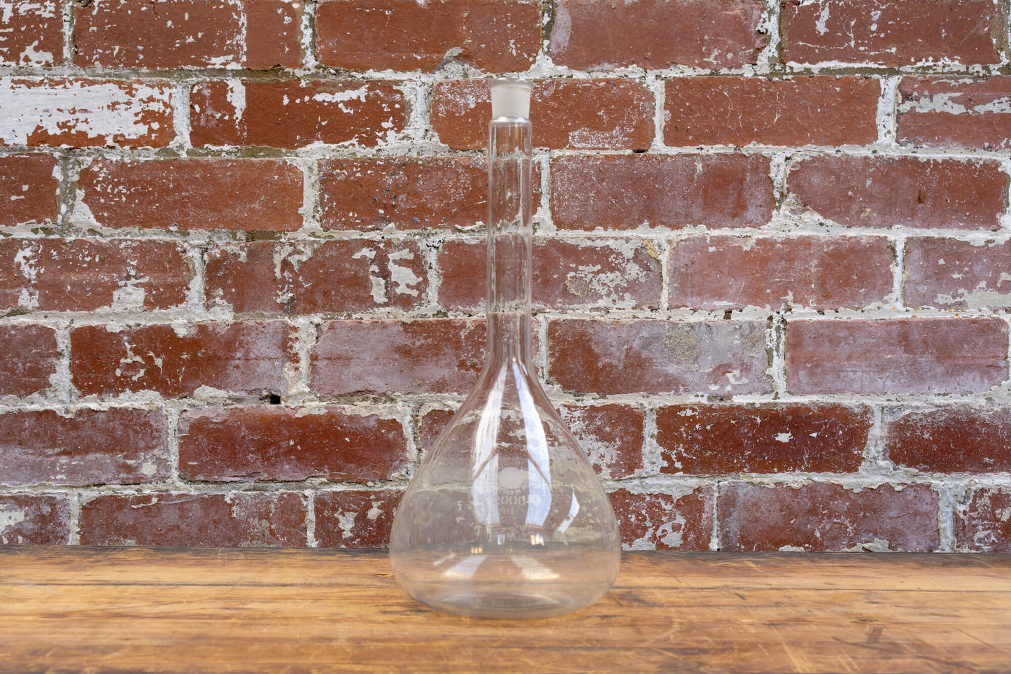 Photo shows a selection of vintage glass laboratory vessels on a table. The background is a red rustic brick wall.