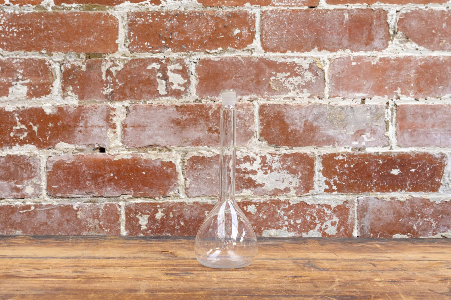 Photo shows a selection of vintage glass laboratory vessels on a table. The background is a red rustic brick wall.