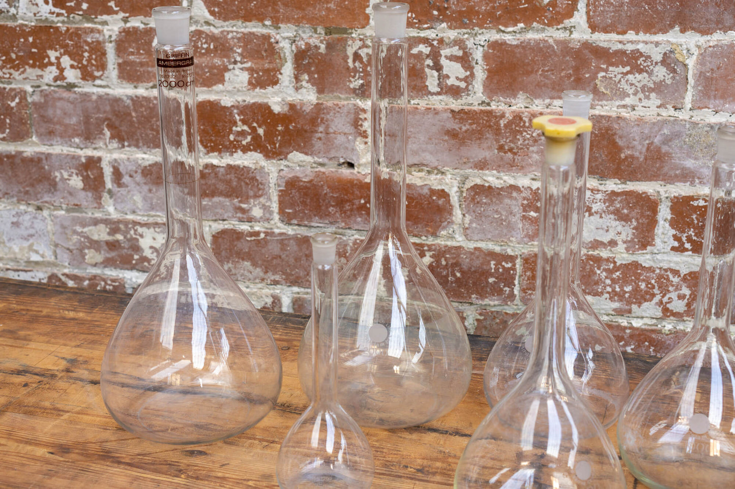 Photo shows a selection of vintage glass laboratory vessels on a table. The background is a red rustic brick wall.