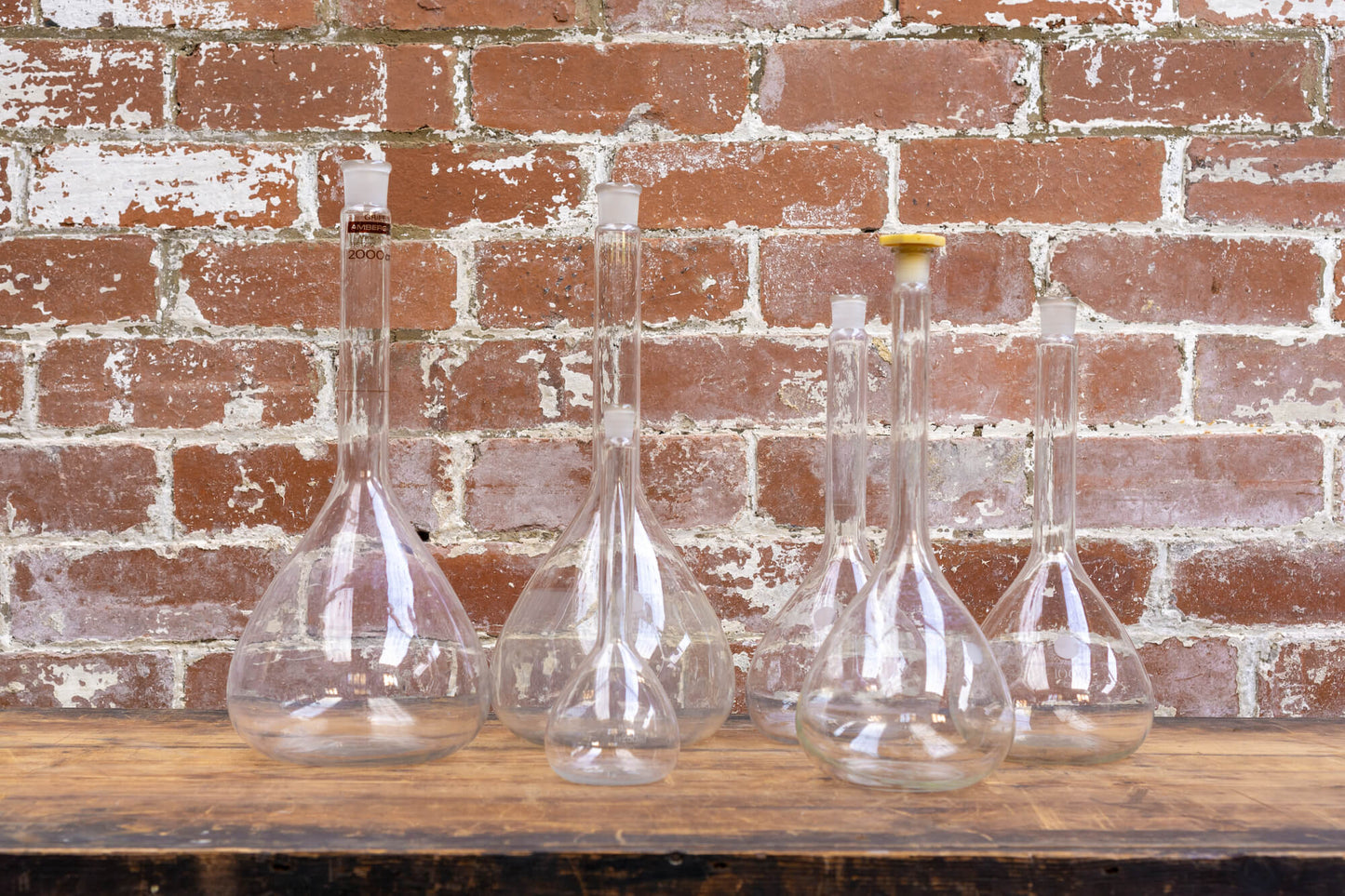 Photo shows a selection of vintage glass laboratory vessels on a table. The background is a red rustic brick wall.