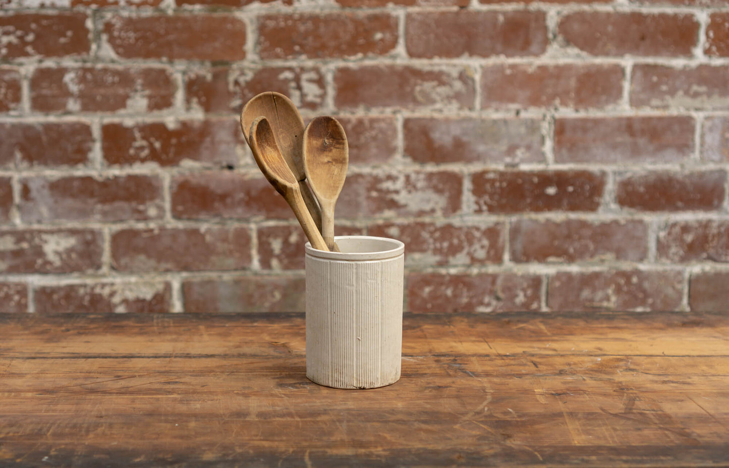 Photo shows a salvaged Victorian stoneware jam conserve jars against a brick wall background. There are wooden spoons inside. The jar is cream coloured with a glaze.