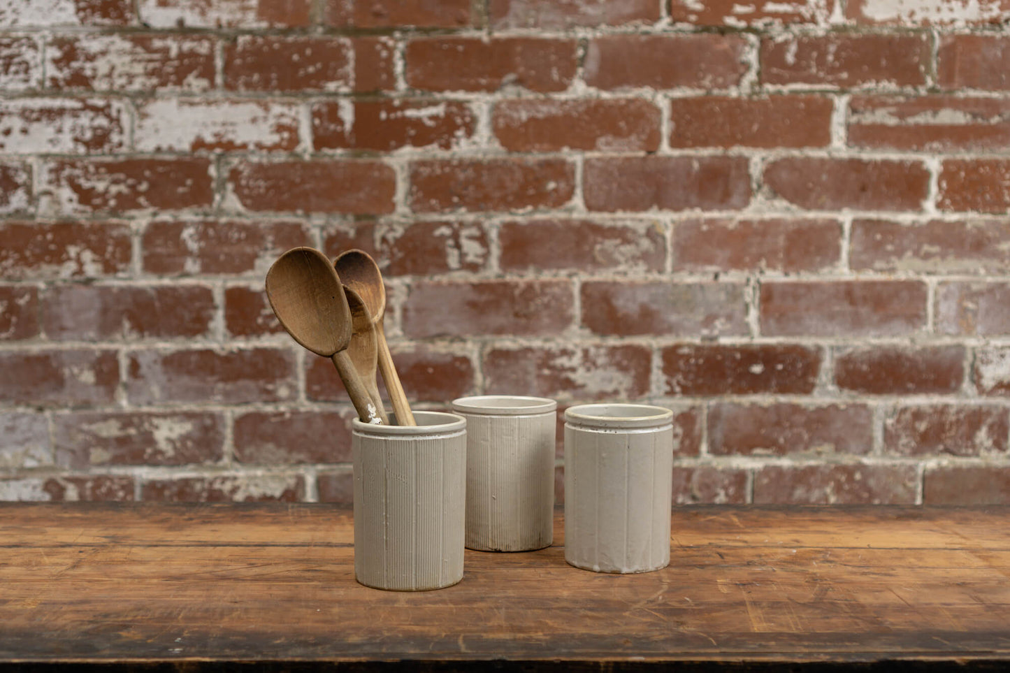 Goods In Victorian Stoneware Conserve Pots JarsPhoto shows 3 various sized salvaged Victorian stoneware jam conserve jars against a brick wall background. The jars are cream coloured with a glaze. One of the jars has vintage wooden spoons stored inside.