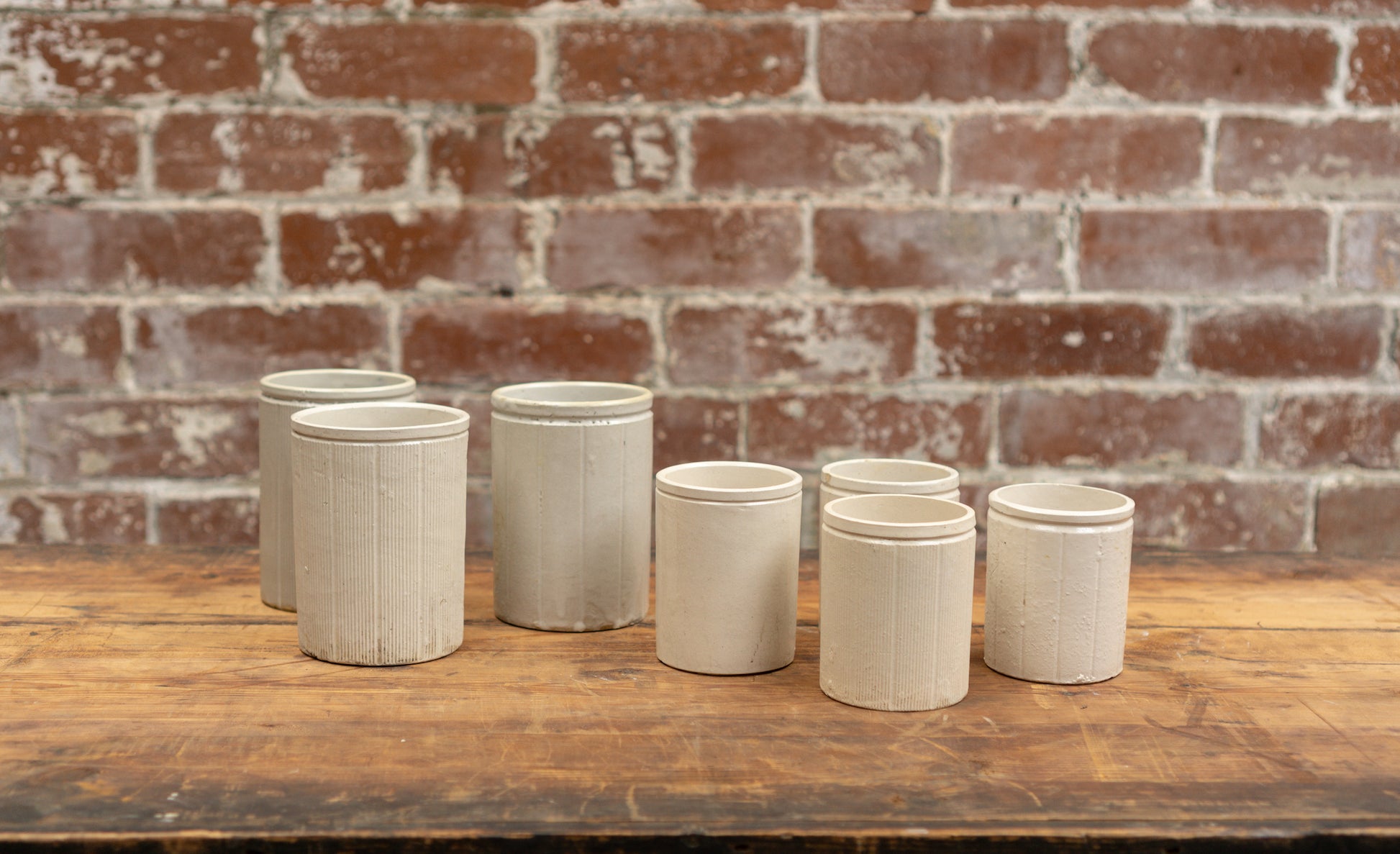 Photo shows 6 various sized salvaged Victorian stoneware jam conserve jars against a brick wall background. The jars are cream coloured with a glaze.