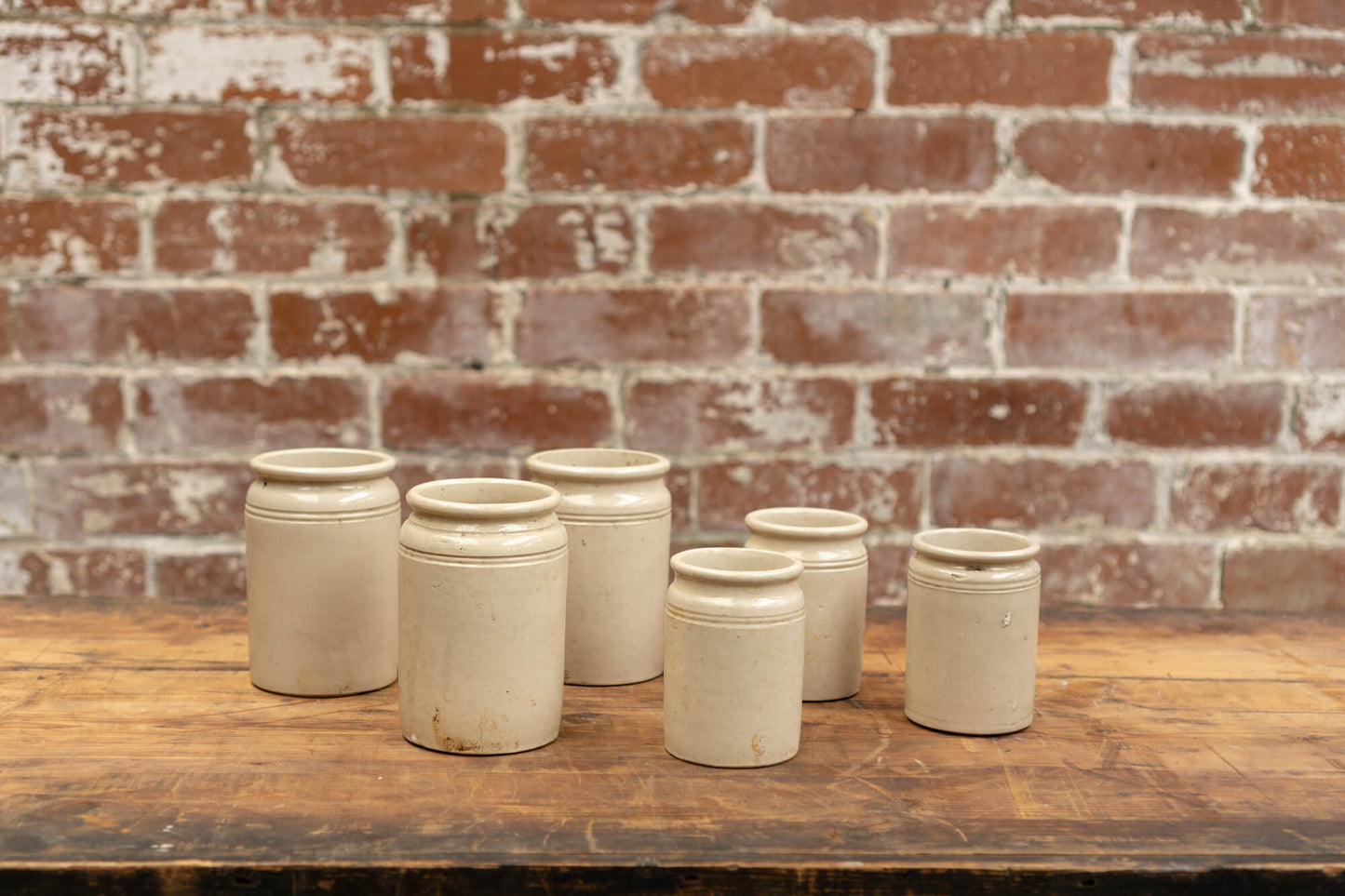 Photo shows 6 various sized salvaged Victorian stoneware jam conserve jars against a brick wall background. The jars are cream coloured with a glaze.