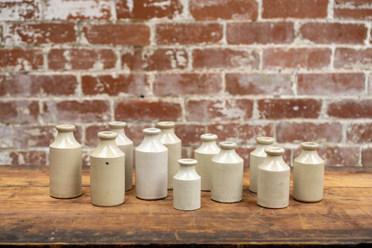 Image shows a collection of victorian stoneware bottles in shades of ivory, displayed neatly against a red brick wall backdrop