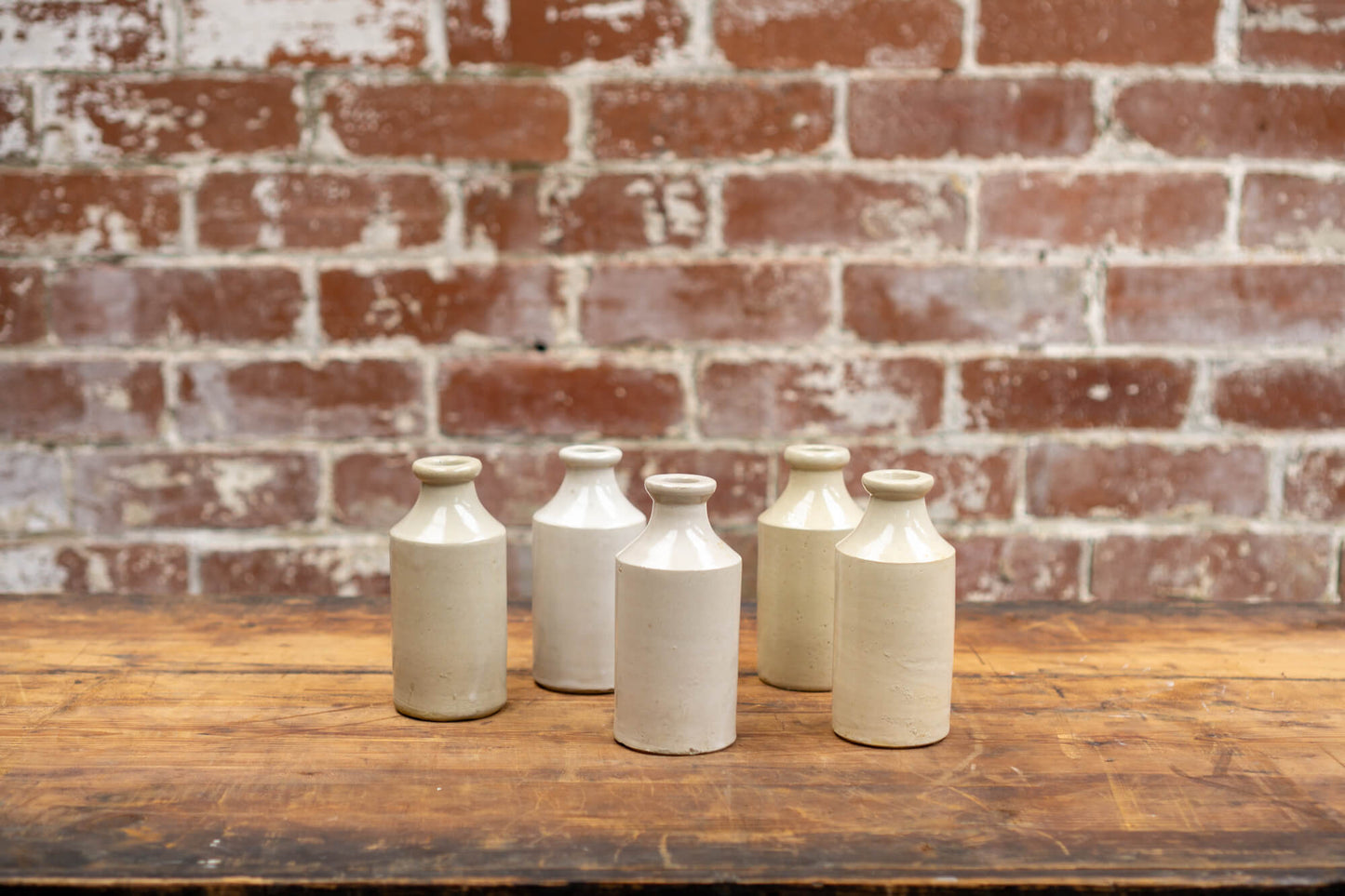 Image shows a collection of victorian stoneware bottles in shades of ivory, displayed neatly against a red brick wall backdrop